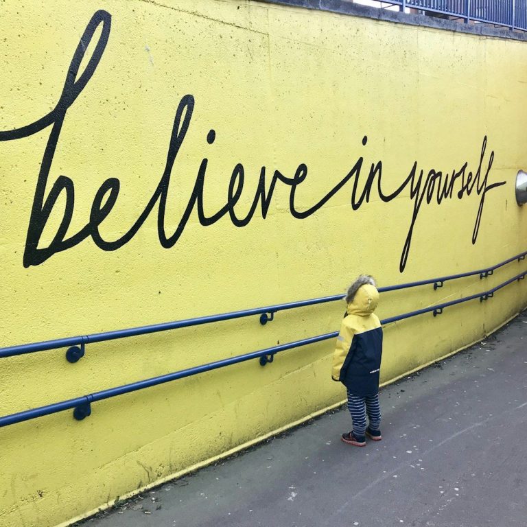 "Believe in yourself" Ein Kind mit gelber Kapuze steht vor einer Wand mit der Aufschrift "Believe in yourself".